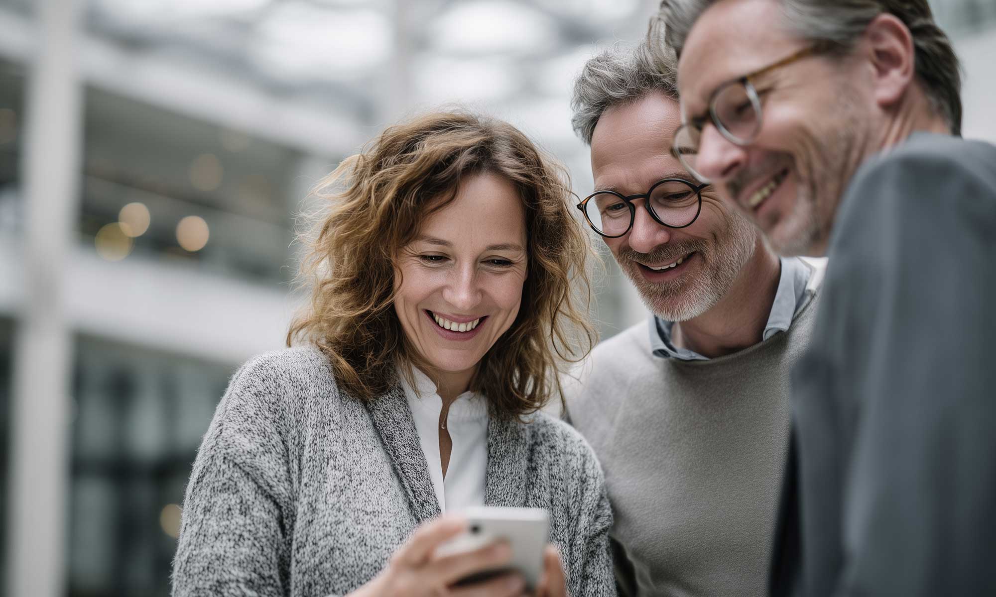 Drei Geschäftspersonen mittleren Alters lächeln, während sie gemeinsam auf ein Smartphone schauen; sie stehen in einem modernen Bürogebäude mit Glasfassade im Hintergrund.